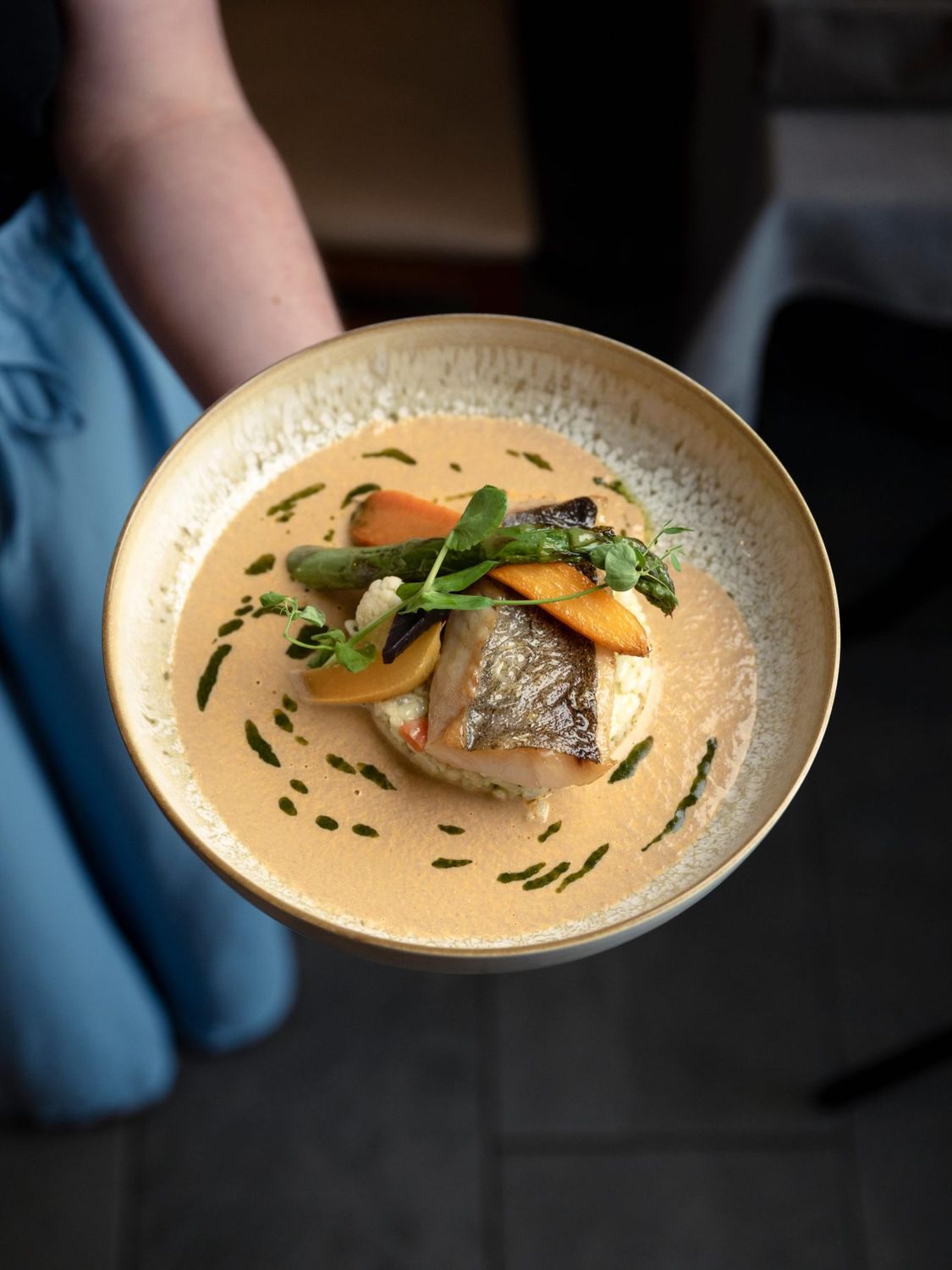 Une assiette de L'Ambroisie présentée à la main, poisson et légumes dans un bouillon crémeux